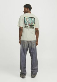 Lys grå grafisk t-shirt med skrækhorrortema tryk på ryggen, der har livlige farver. Sættes sammen med løstsiddende mørkeblå denimbukser.