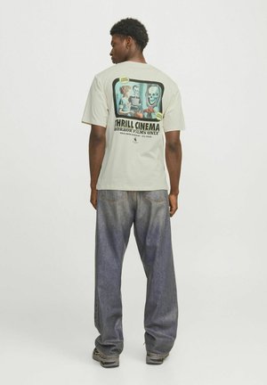 Lys grå grafisk t-shirt med skrækhorrortema tryk på ryggen, der har livlige farver. Sættes sammen med løstsiddende mørkeblå denimbukser.