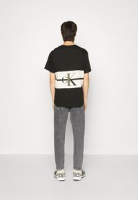 Muž stojící zády, oblečený v černobílém tričku Calvin Klein, šedých džínách a bílých teniskách na jednobarevném pozadí.