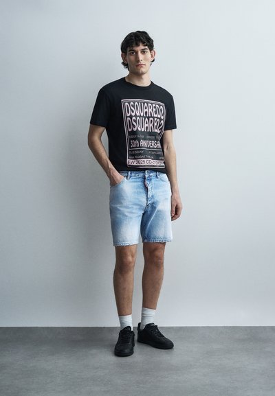 Jeune homme debout contre un mur uni, portant un t-shirt noir à motif, un short en jean clair, des chaussettes blanches et des baskets noires, mains dans les poches.