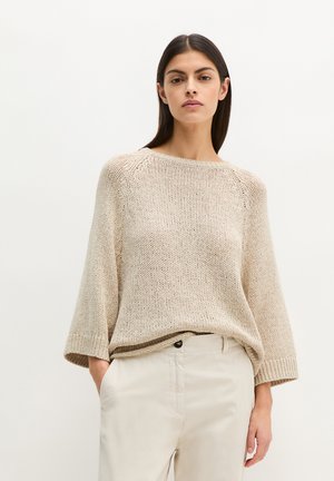 Kobieta o długich ciemnych włosach, ubrana w luźny beżowy sweter z dzianiny i jasnobeżowe spodnie, z rękami w kieszeniach, stojąca na jednolitym tle.