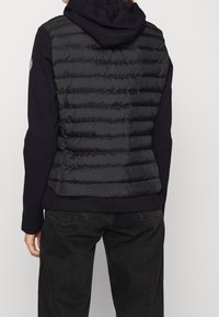 Gilet imbottito nero con cuciture orizzontali, abbinato a una felpa scura sotto. La texture è liscia con dettagli trapuntati. Design elegante.