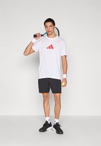 Fehér sportpóló piros Adidas logóval, fekete rövidnadrág narancssárga díszítéssel, teniszütőt és labdát tart, fekete tornacipőt visel.