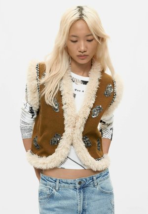 Junge Frau mit langen blonden Haaren, die eine braune, mit Pelz besetzte Weste über einem weißen, bedruckten Langarmshirt und hellblauen Jeans trägt.