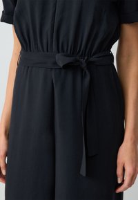 Combinaison noire marine avec une taille cintrée, dotée d'une ceinture nouée, de manches courtes et d'un tissu léger et lisse qui tombe doucement.