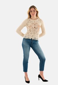 Cardigan in pizzo crema con motivi intricati, maniche lunghe e bottoni frontali, abbinato a jeans slim-fit blu e scarpe con tacco nere.