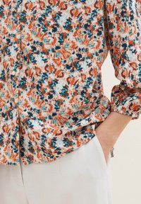 Gros plan sur une personne portant un chemisier floral multicolore à boutons, une main posée négligemment dans la poche d'un pantalon beige clair.