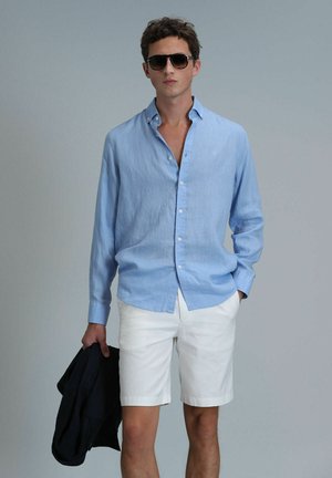 Lichtblauw linnen shirt met lange mouwen, met een button-down kraag, gecombineerd met witte shorts. Houdt een donker jasje vast, draagt een zonnebril. Minimale details.