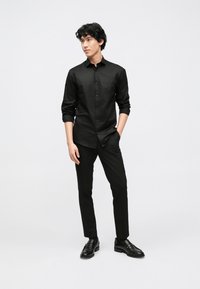 Zwart overhemd met knopen, met een gladde textuur, gecombineerd met strakke zwarte broek en zwarte nette schoenen, weergegeven tegen een witte achtergrond.