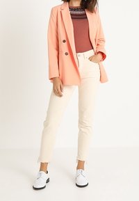 Blazer en pêche avec des boutons doubles, haut bordeaux, et jean crème avec des ourlets effilochés ; complété par des chaussures blanches avec des semelles noires.