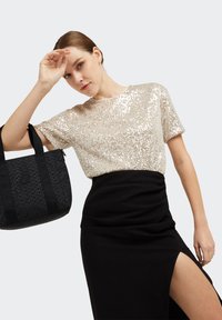 Short-sleeve, metallic gold sequin top paired with a black, knee-length skirt featuring a side slit. Black tote bag with patterned design.