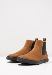 Brązowe zamszowe botki za kostkę z elastycznymi bocznymi panelami, czarnymi akcentami i czarną gumową podeszwą. Szwy dekoracyjne wokół krawędzi.