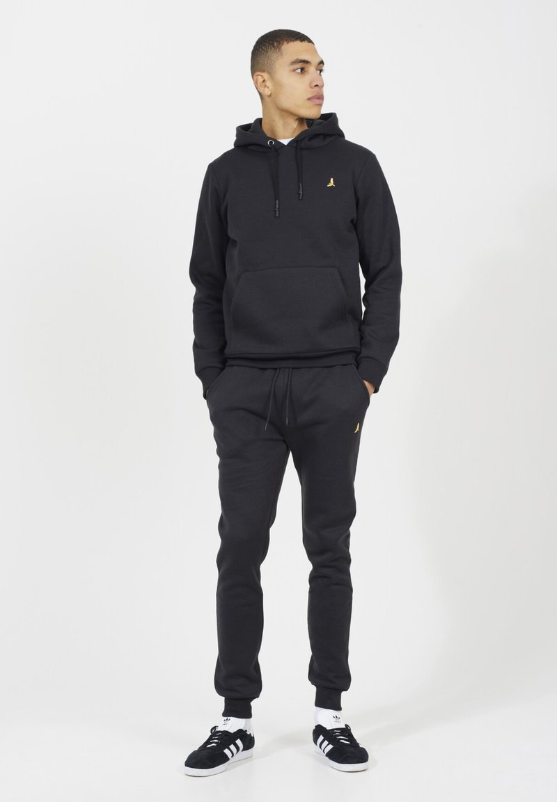 Felpa nera in cotone con tasca a marsupio sul davanti e logo dorato. Abbinata a pantaloni coordinati e sneakers nere con dettagli bianchi.