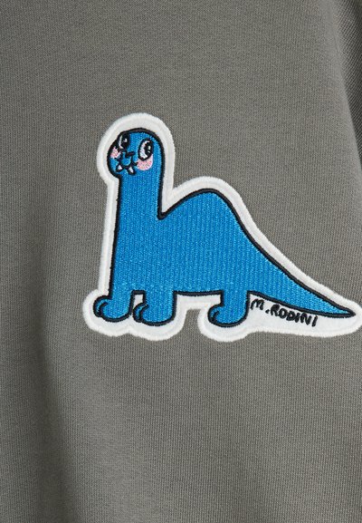 Tejido gris con un diseño de dinosaurio bordado en azul con contorno blanco, pequeños detalles en negro y la firma "M. Rodini" en la parte inferior.