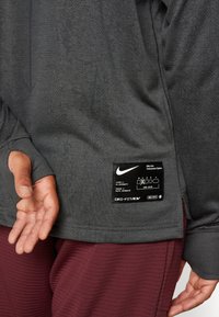 Persona con camiseta de manga larga Nike Dri-FIT de color gris oscuro con agujero para el pulgar y pantalones burdeos texturizados, mostrando la etiqueta de Nike en el dobladillo de la camiseta.