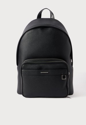 Crni ruksak s glatkom teksturom, zaobljenim vrhom, prednjim džepom na patentni zatvarač i metalnim dodacima. Ima čvrstu ručku na vrhu za lako nošenje.