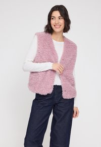 Flauschige pinke Weste mit offenem Design und ohne Verschlüsse, getragen über einem weißen Langarmshirt, kombiniert mit dunkelblauen weit geschnittenen Hosen.