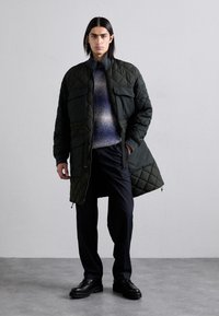 Manteau matelassé vert foncé avec un col haut, deux poches avant et une coupe mi-longue, associé à un chandail dégradé bleu et un pantalon noir.