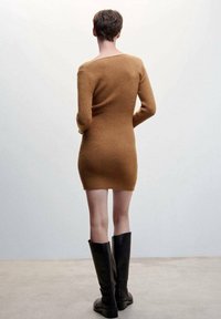 Robe marron en maille côtelée avec manches longues, silhouette ajustée et large encolure. Associée à des bottes hautes noires, texture cuir lisse.