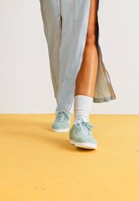 Zapatillas de ante verde menta con suelas y cordones blancos, usadas con calcetines deportivos blancos, en un conjunto de denim claro sobre un fondo amarillo.
