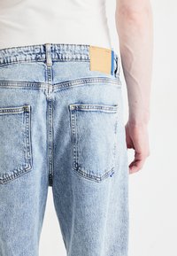 Pantalones vaqueros de denim azul claro con corte recto, que cuentan con una parche de cuero y dos bolsillos traseros. La tela presenta una ligera textura.