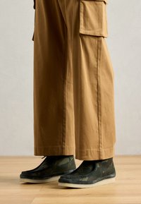 Kamelan mönstrade ankelskor i mörkgrön med en grå sula; kombineras med vida beige byxor med sidofickor. Bakgrund av trägolv.