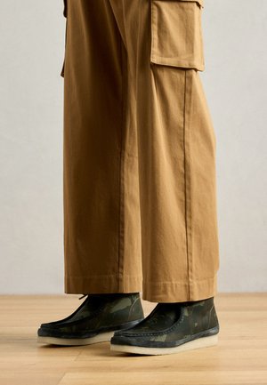 Camouflage-patroon enkellaarzen in donkergroen met een grijze zool; gecombineerd met ruime beige broek met zijzakken. Houten vloer op de achtergrond.