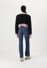 Svart fluffig croppad tröja med randig nederkant i röd och vit, kombinerad med blå utsvängda jeans och beige sneakers. Visas från baksidan.
