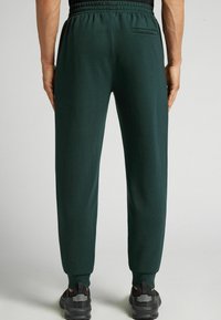 Donkergroene joggers met een elastische tailleband en taps toelopende benen, voorzien van een achterzak en een zachte stoftextuur. Draag met zwarte sneakers.