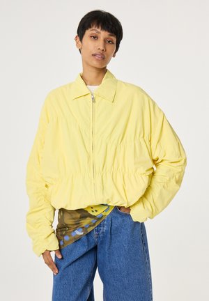 Person wearing a loose yellow zip-up jacket over a patterned shirt, paired with blue denim jeans, standing with one hand in pocket.