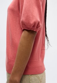 Közelkép egy személy karjáról, aki korallszínű rövid ujjú pulóvert és bézs nadrágot visel, az oldalán látható fonott frizurával.