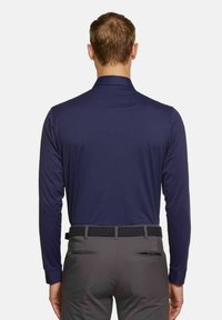 Marineblaues Langarmhemd mit Kragen, aus glattem Stoff, kombiniert mit grauen Hosen, die über einen geflochtenen schwarzen Gürtel und einen Reißverschluss in den Gesäßtaschen verfügen.