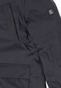 Tamsiai mėlynas pagalvotas švarkas su lygiu paviršiumi, pečių siūlės detale ir kvadratiniu logotipo ženkleliu. Šone esančio kišenė su elegantišku atidarymu.