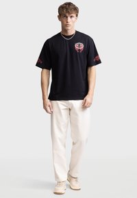 Camiseta de algodón negra con diseños gráficos rojos en el pecho y la manga, combinada con pantalones claros y zapatillas beige.