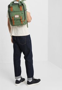 Zelený látkový batoh s hnědými koženými detaily, obdélníkový tvar, přední kapsa na zip a dvojité horní úchyty, nošený na zádech osoby.