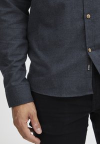 Dunkelgrauhe Button-up-Shirt aus weichem Stoff, mit Holzknöpfen, abgerundetem Saum und einem Etikett an der Seite, kombiniert mit schwarzen Hosen.