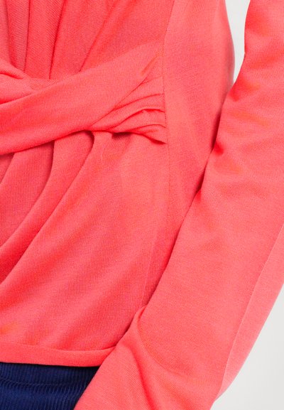 Top lungo a maniche con nodo in vita di colore corallo. Tessuto testurizzato con finitura liscia. Presenta una leggera costina e maniche sartoriali.