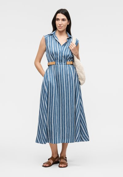 Γυναίκα με αμάνικο μπλε και λευκό midi φόρεμα με κάθετες ρίγες και ταμπά ζώνη, κρατώντας μια μπεζ τσάντα ώμου με υφή, φορά καφέ σανδάλια.