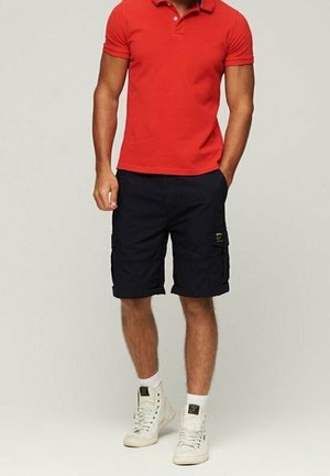 Homme portant un polo rouge, un short cargo noir, des baskets blanches montantes et des chaussettes blanches, debout avec une main dans la poche.