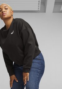 Felpa nera con collo a giro e polsini a costine, con un piccolo logo Puma bianco sul lato sinistro del petto. Indossata con jeans blu.