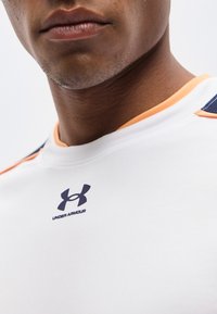 Κοντινό πλάνο του κάτω μέρους του προσώπου και του λαιμού ενός άνδρα που φοράει λευκό αθλητικό πουκάμισο Under Armour με πορτοκαλί και ναυτικά λεπτομέρειες.