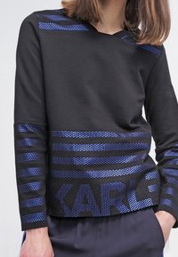 Polo de manga larga negro con acentos de rayas azules. Presenta un tejido texturizado en la mitad inferior y letras impresas. Diseño de escote en V.