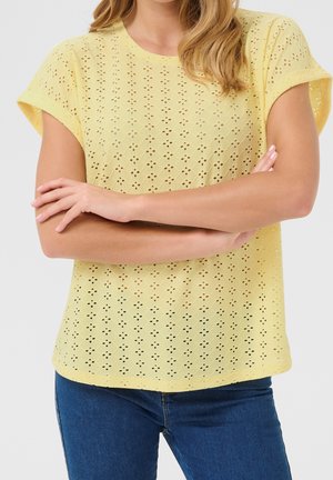 Kobieta ze skrzyżowanymi ramionami, ubrana w żółtą bluzkę z krótkim rękawem z haftowanym wzorem oraz niebieskie dżinsy, na jednolitym tle.