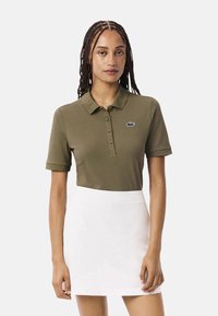 Polo de color verde oliva con mangas cortas y una tapeta de botones, con un pequeño logotipo de Lacoste. Combinado con una falda blanca.