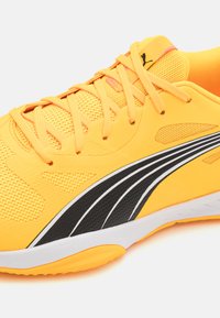 Scarpa sportiva gialla con tomaia in rete, dettagli a strisce nere e bianche e lacci piatti, con suola in gomma testurizzata.