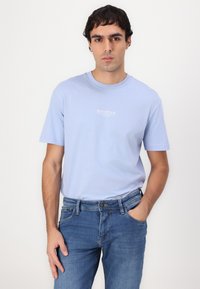 T-shirt en coton bleu clair à manches courtes et col rond, arborant un petit logo blanc sur la poitrine. Porté avec un jean en denim bleu.