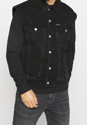 Hombre con chaqueta vaquera negra sin mangas sobre camisa negra de manga larga y vaqueros gris oscuro, con botones plateados en la chaqueta.