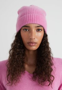 Bonnet rose en matière douce côtelée, avec un design ample. Porté avec un pull rose assorti.
