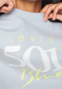 Mains ajustant le col d'un sweat-shirt bleu clair avec le texte jaune et blanc "Levi's Soul Bl" sur le devant.
