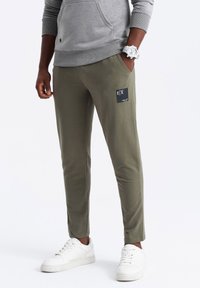 Olivegrüne Jogginghosen aus weichem Material mit schmaler Passform, ausgestattet mit einem schwarzen Logo-Patch auf der linken Tasche und einem elastischen Bund. Kombiniert mit weißen Sneakers.